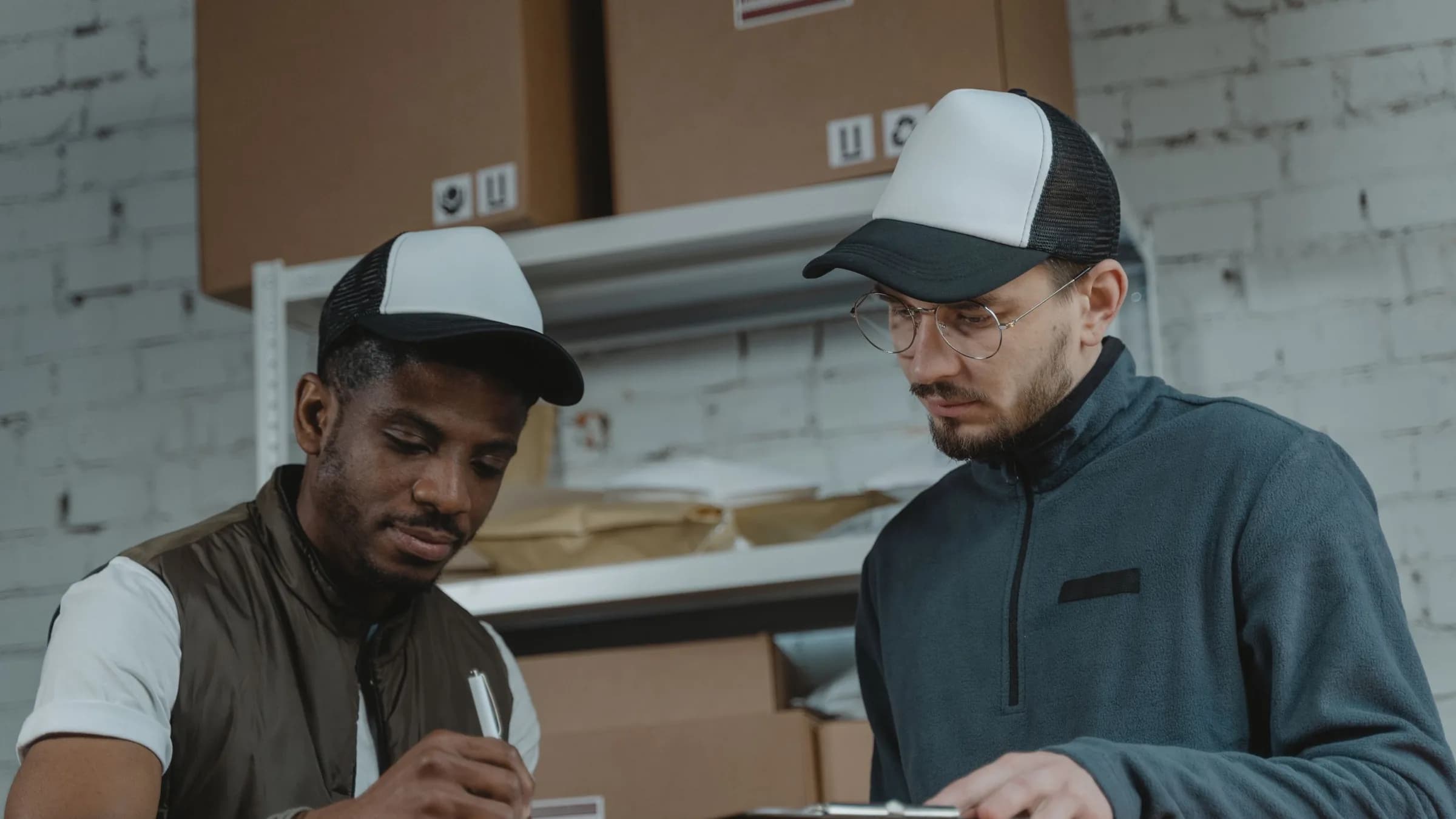 Two warehouse staff in caps go over paperwork together in front of shelves of boxes.