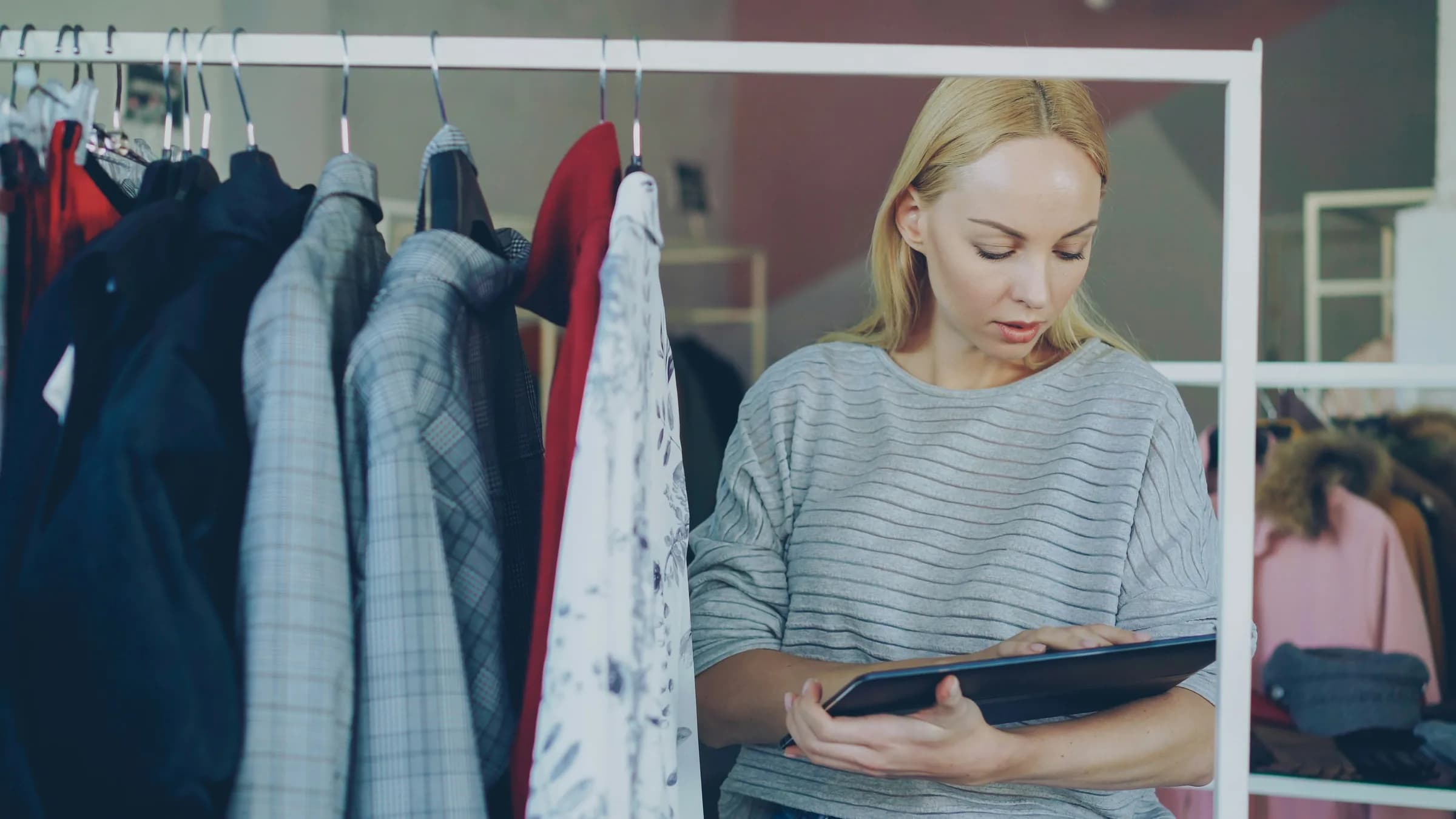 A shop owner checks stock on a tablet beside a rail of jackets and shirts.