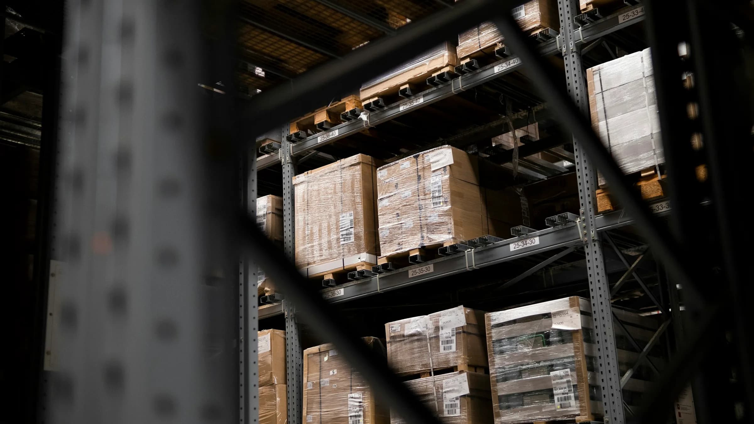 Cartons of stock stacked on pallets on tall metal racking, seen through the frame of the shelving.
