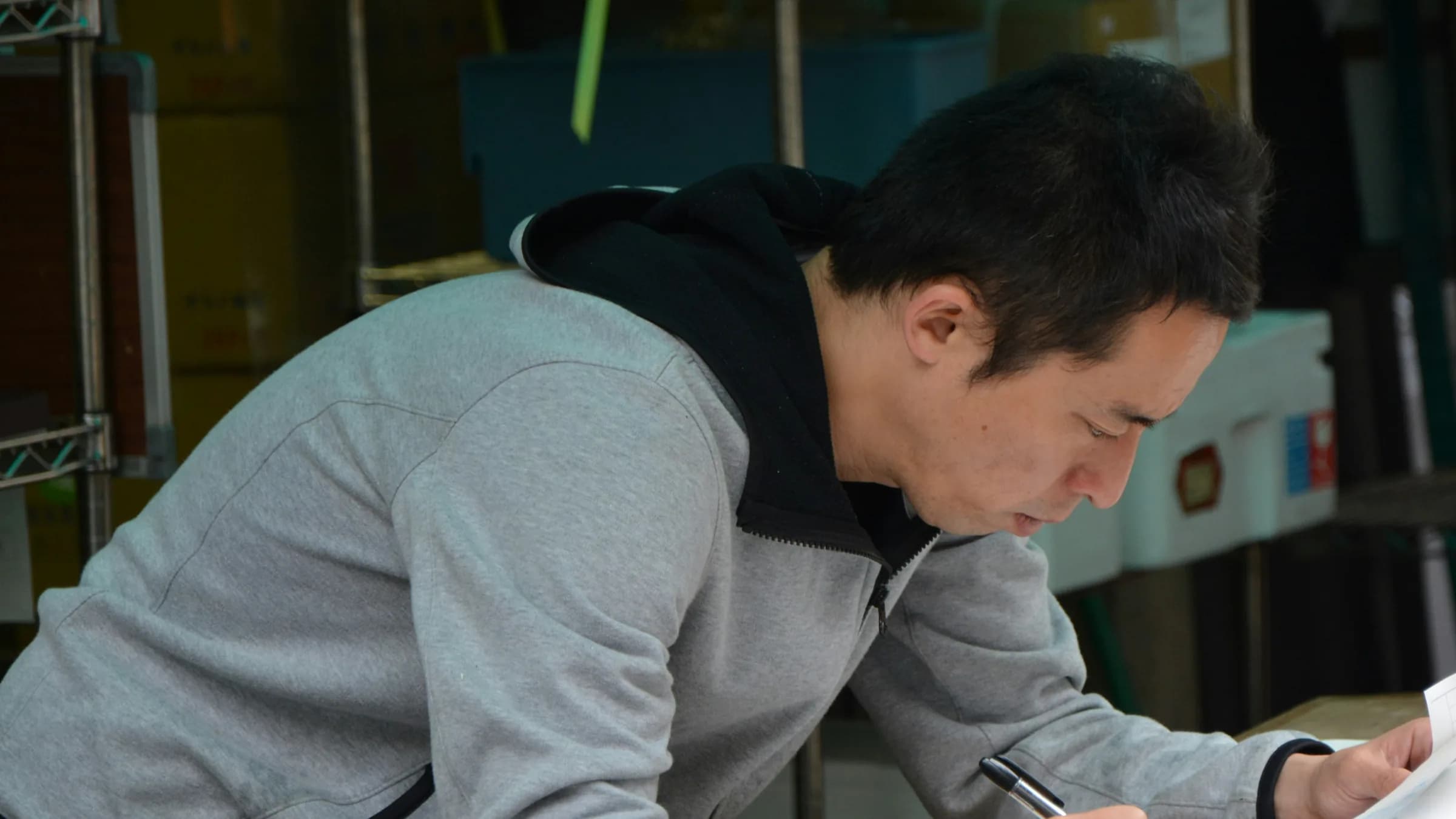 A man in a grey hoodie leans over a stockroom bench, writing figures on a sheet of paper.