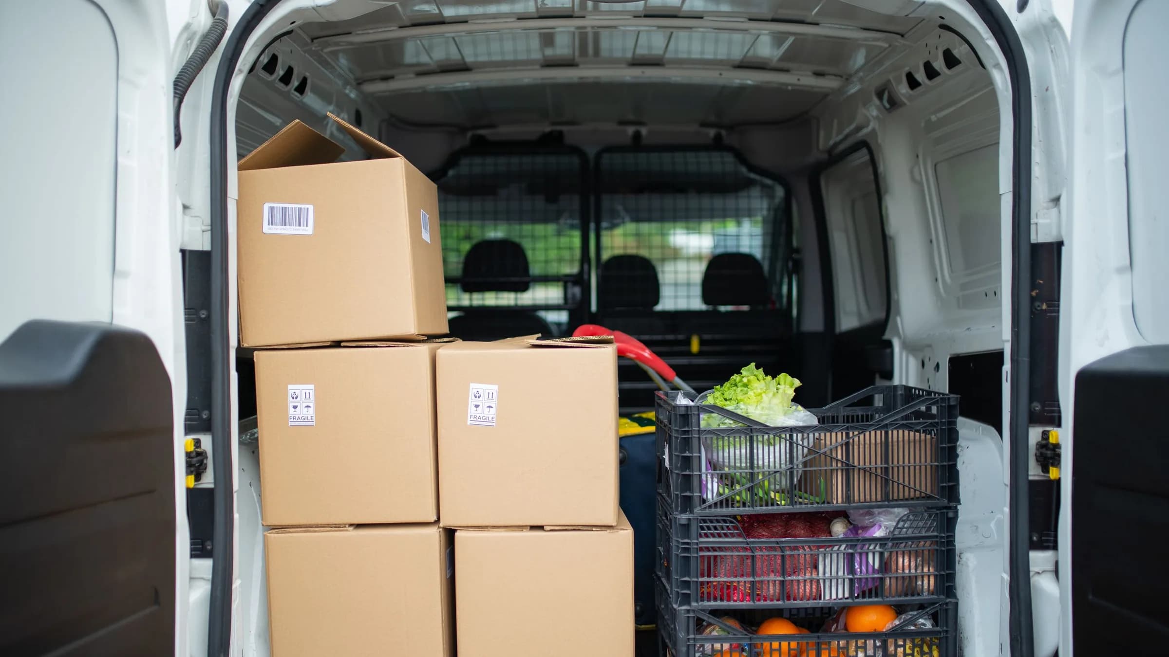 The open back of a delivery van loaded with cartons and a crate of fresh fruit and vegetables.