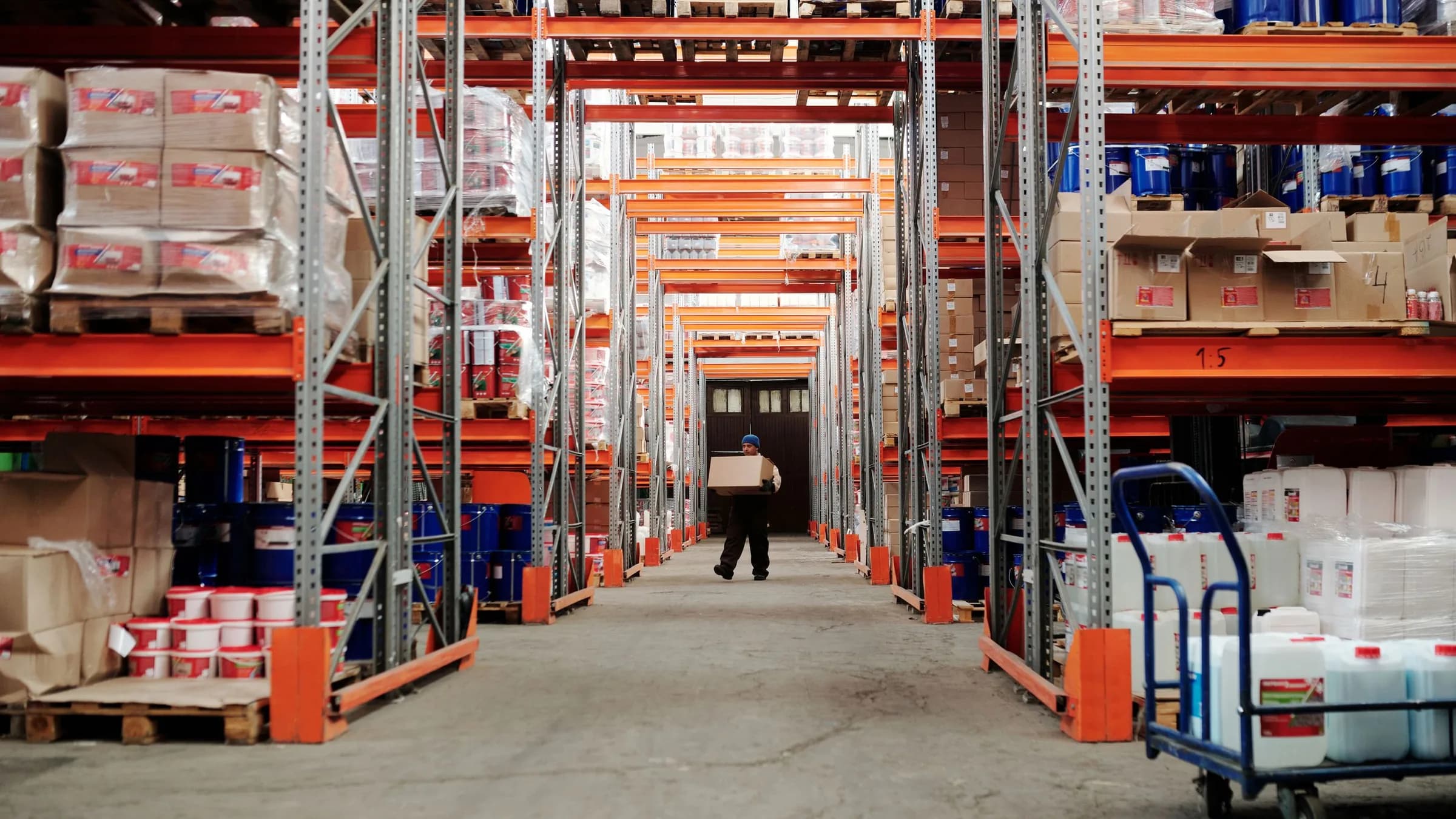 A worker carries a carton down a long warehouse aisle lined with pallet racking full of stock.